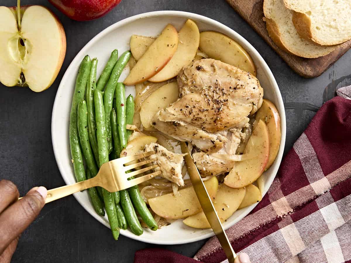 Overhead view of chicken and apples skillet dinner being cut into with a knife and fork on a plate with green beans.