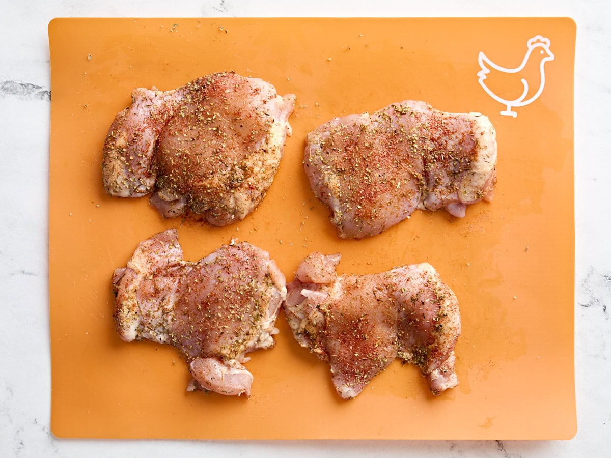 Chicken thighs seasoned with a Greek seasoning mix on a meat preparation mat.