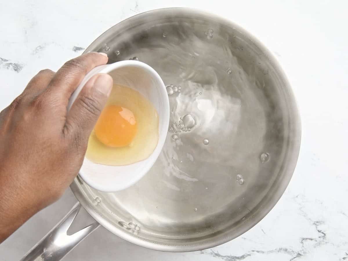 A cracked egg being poured into a boiling pan of water to make poached eggs.