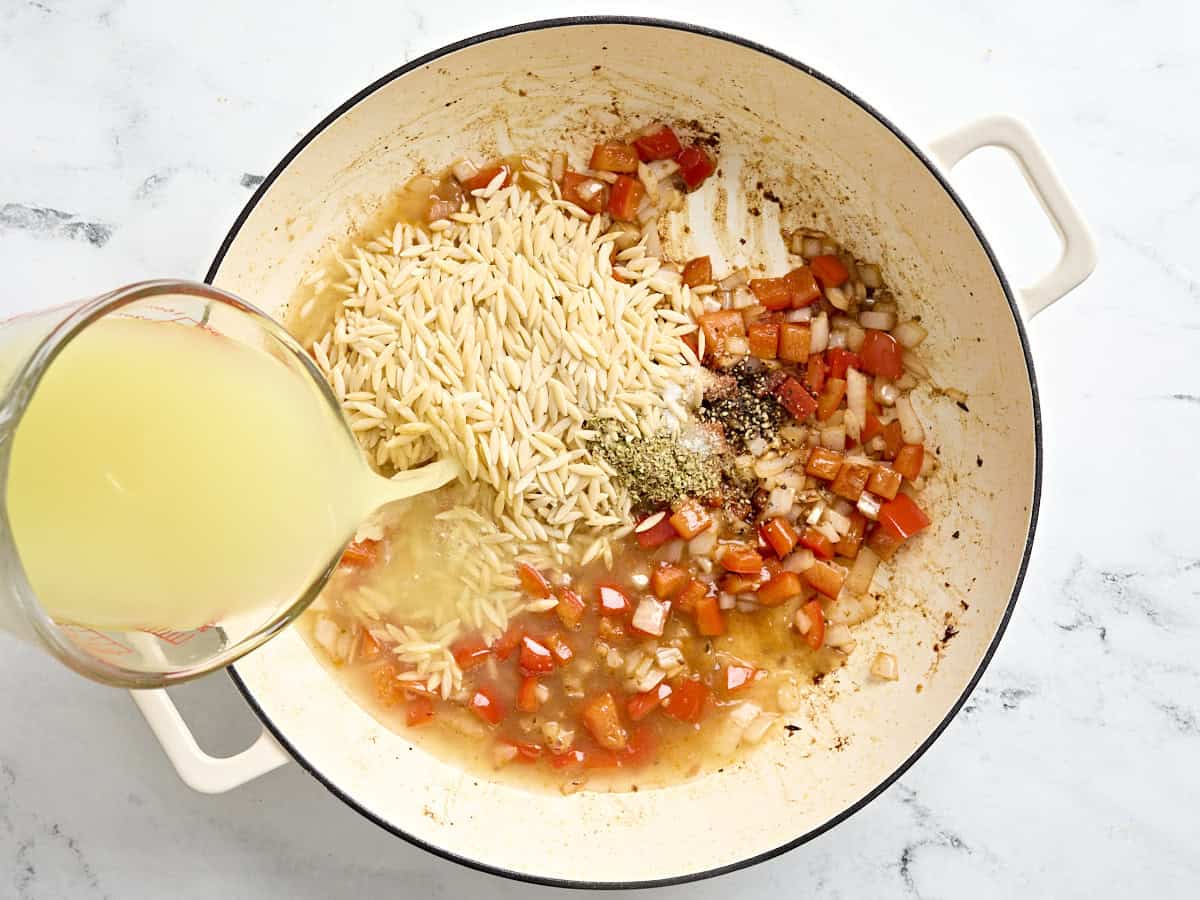 Chicken broth being pored into a skillet with diced vegetables and dried orzo pasta.