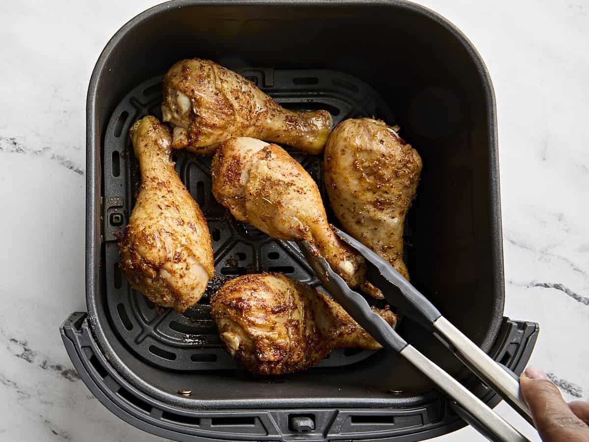 Chicken drumsticks cooking in an air fryer, with tong flipping one over halfway through cooking.