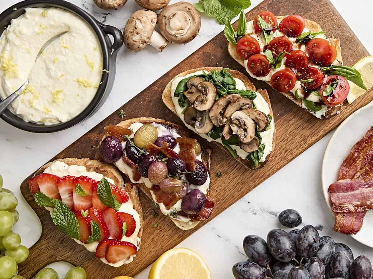 Ricotta toast 4 ways on a wooden serving platter, one topped with strawberries and mint, the next with roasted grapes and bacon, another with blistered tomatoes and basil, and the last with mushrooms and wilted spinach.