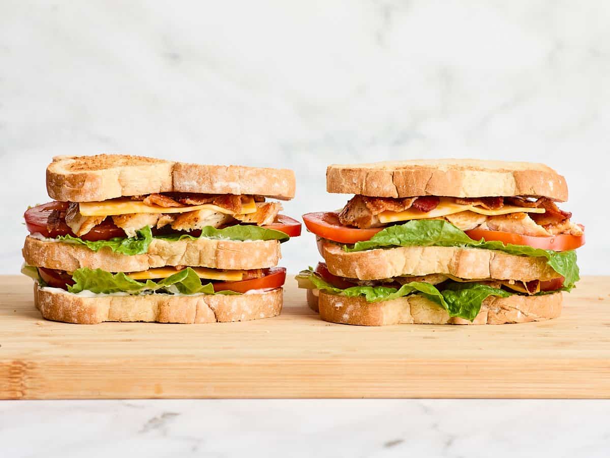 Side view of two chicken club sandwiches on a wooden cutting board.