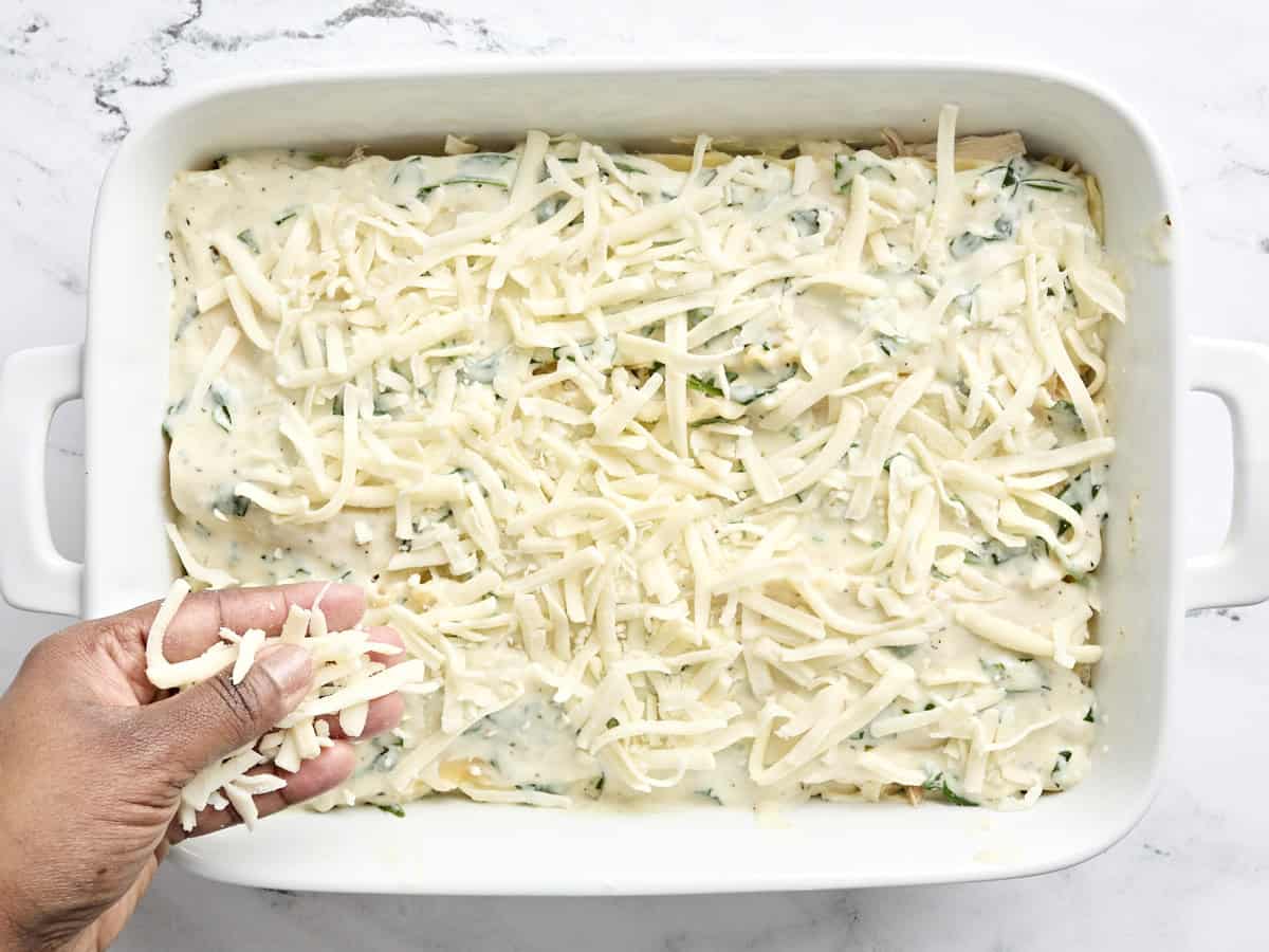 A hand sprinkling cheese over white chicken lasagna in a baking dish.