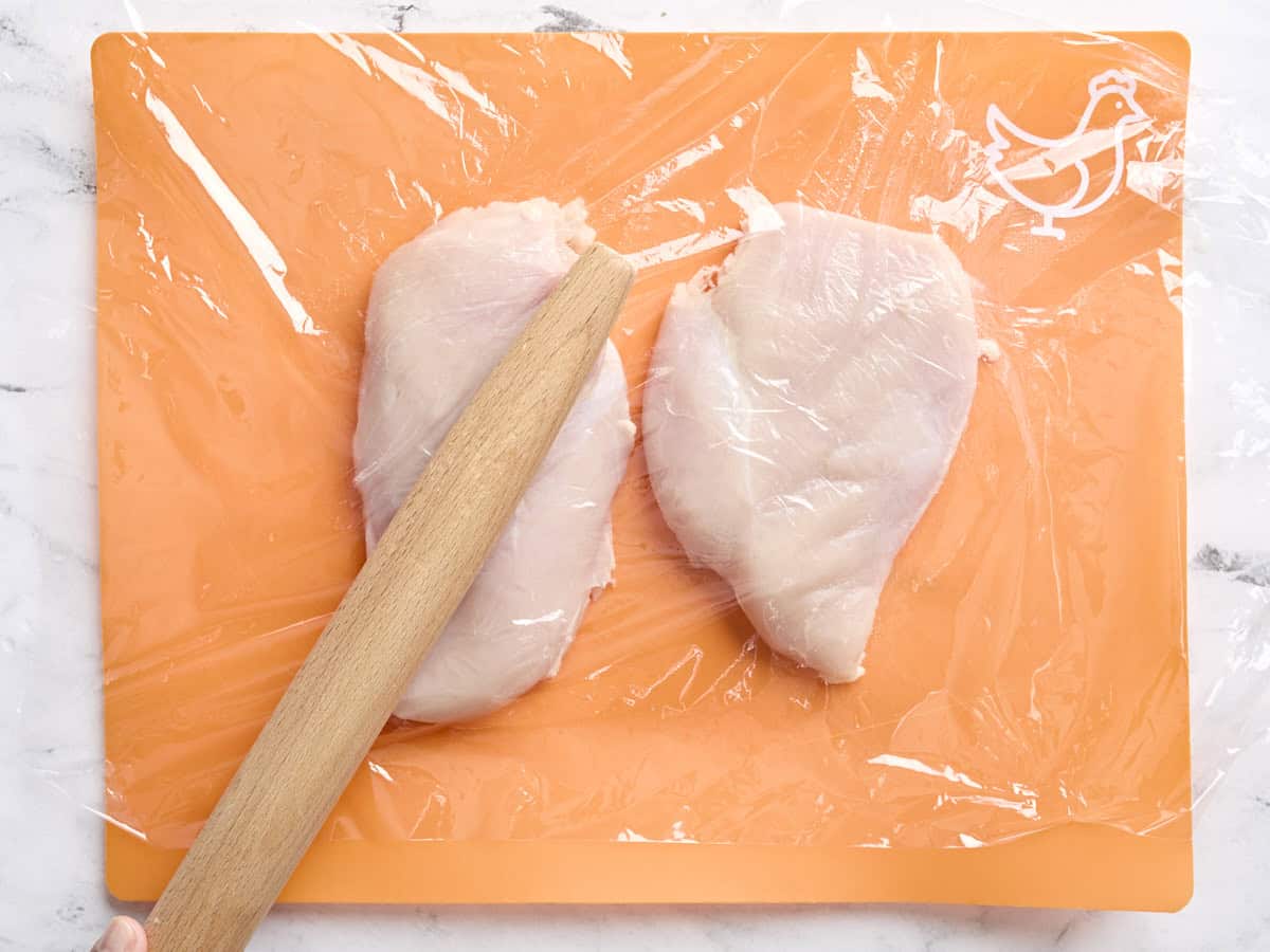 Two chicken breasts on a cutting board covered with plastic wrap, being pounded with a rolling pin.