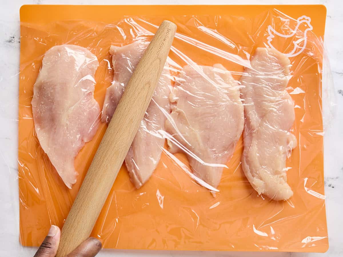 Chicken breasts, covered with plastic wrap, being pounded with a rolling pin.