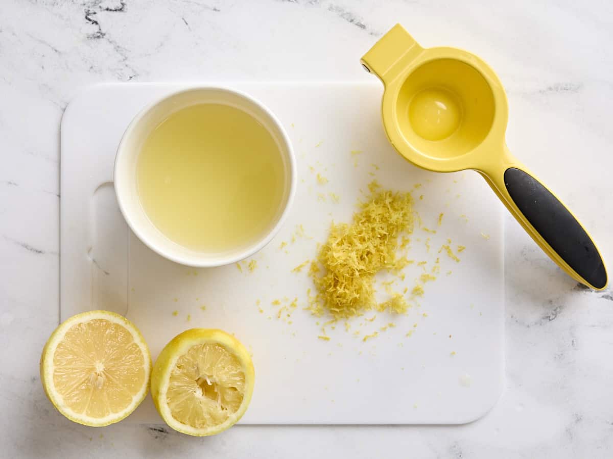 Two lemon halves on a white cutting board, with a hand-held juicer, lemon zest, and a bowl of lemon juice.