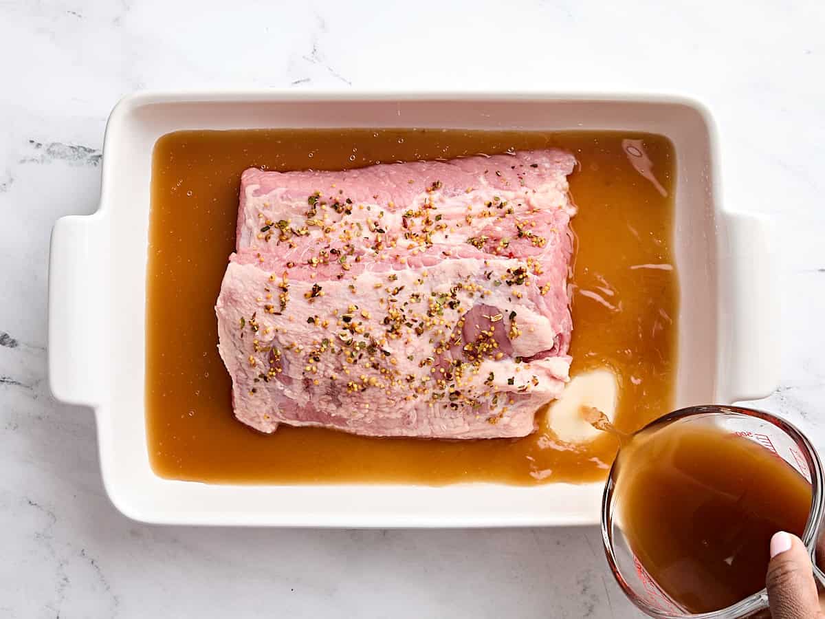 Corned beef topped with seasonings in a baking dish with beef broth being poured into the baking dish.