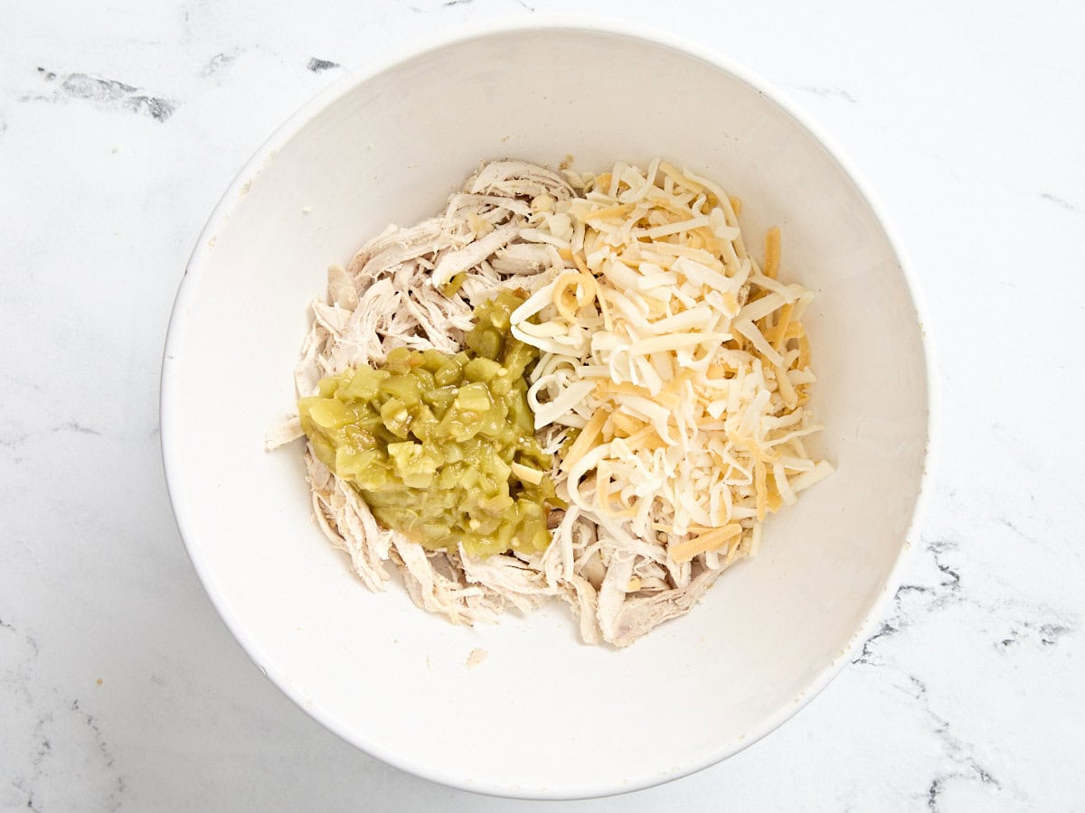 Diced green chilis and shredded cheese added to a bowl of seasoned shredded chicken.