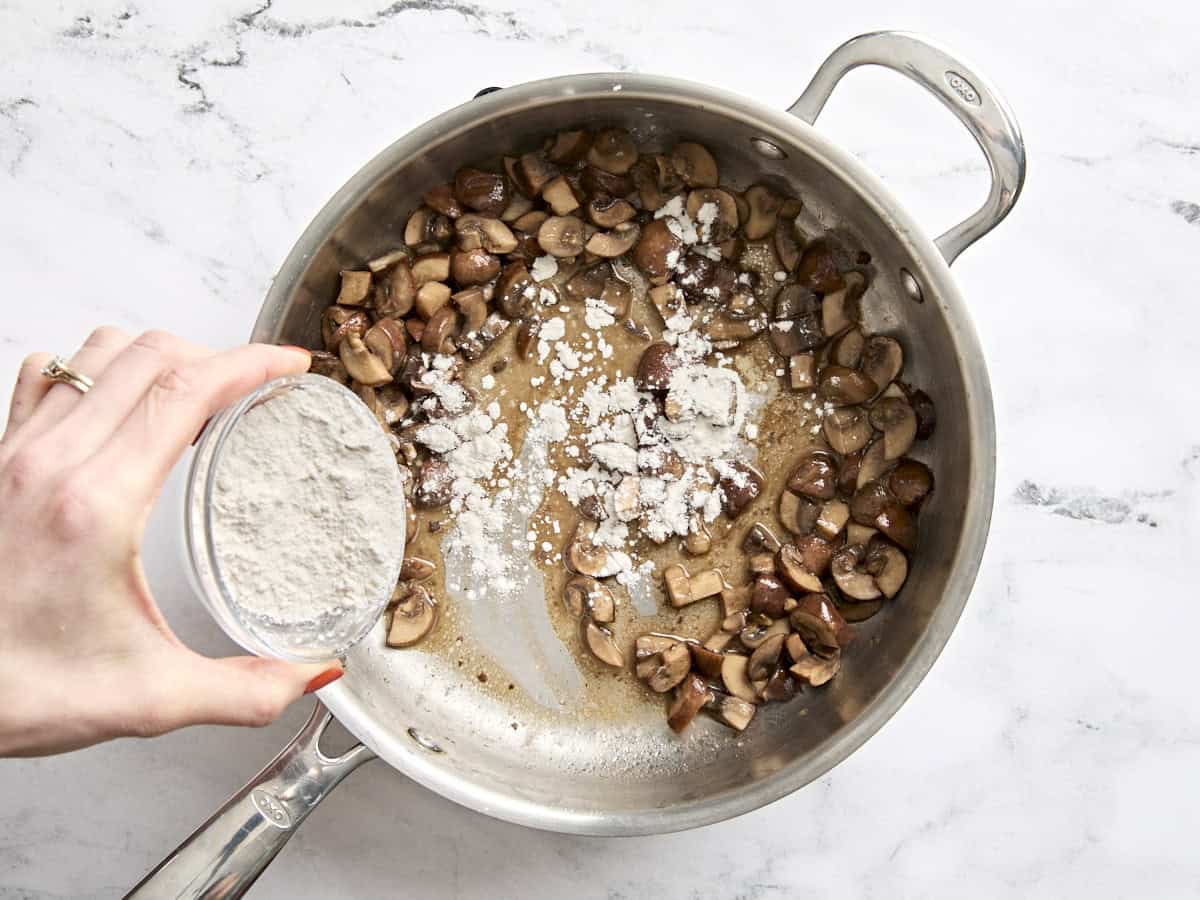 Flour being sprinkled into a saute pan with diced mushrooms and melted butter.