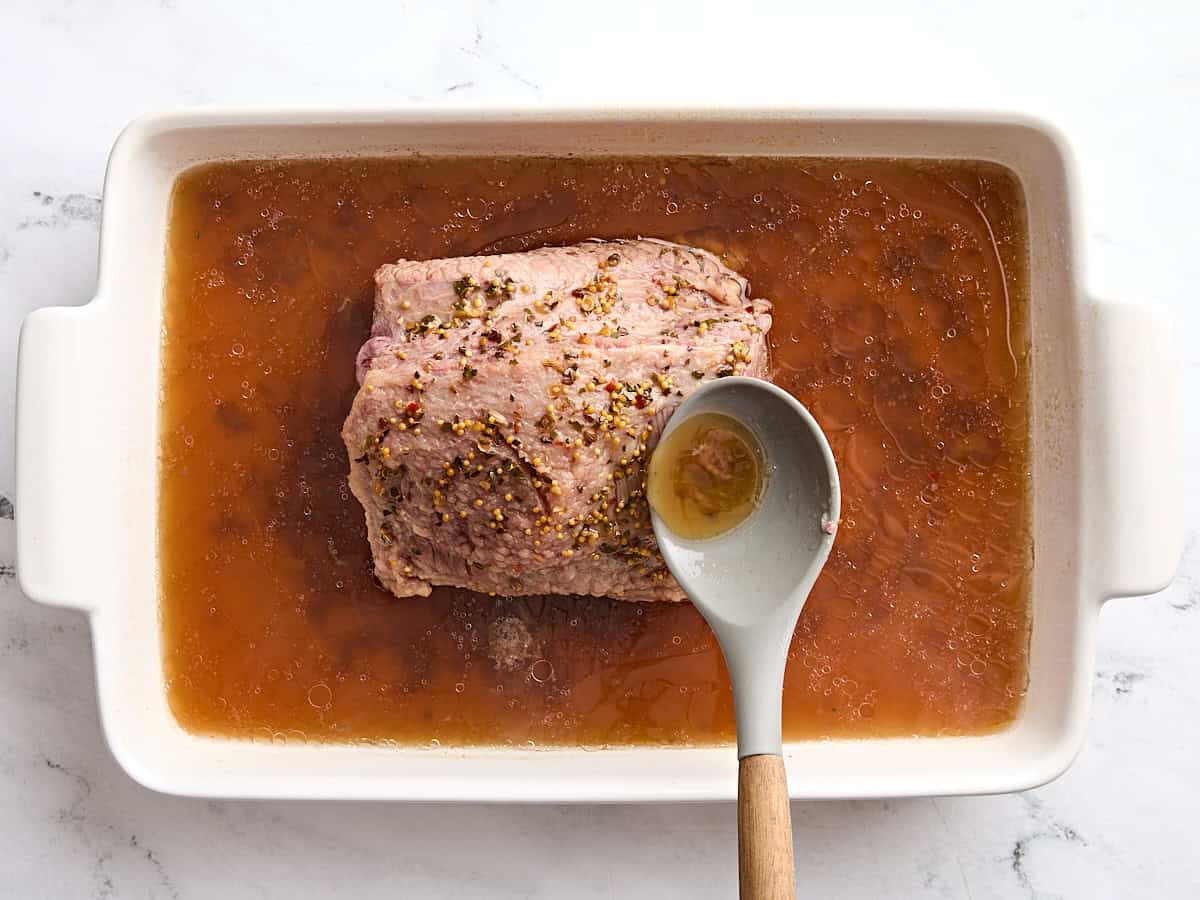 A spoon basting a piece of corned beef in a baking dish.