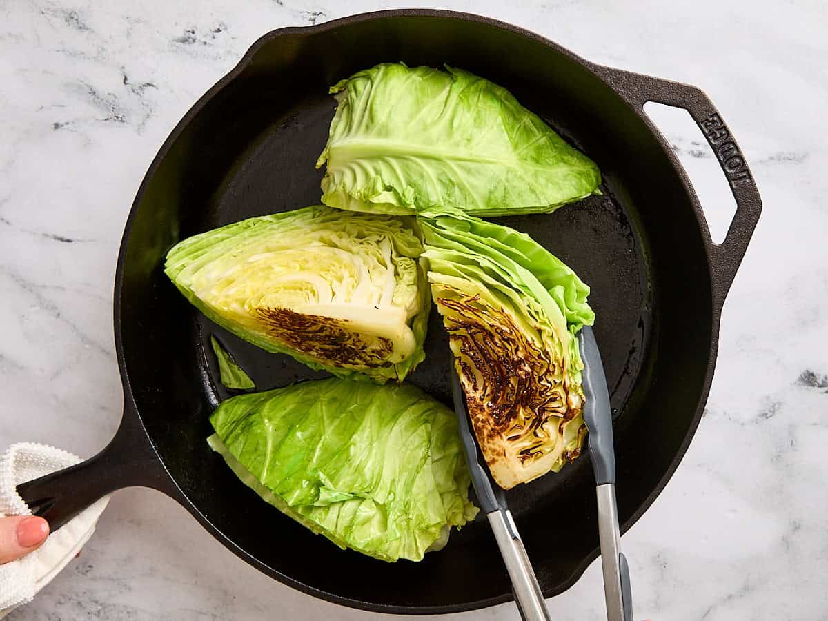 Kitchen tongs flipping over cabbage steaks in a cast iron skillet.