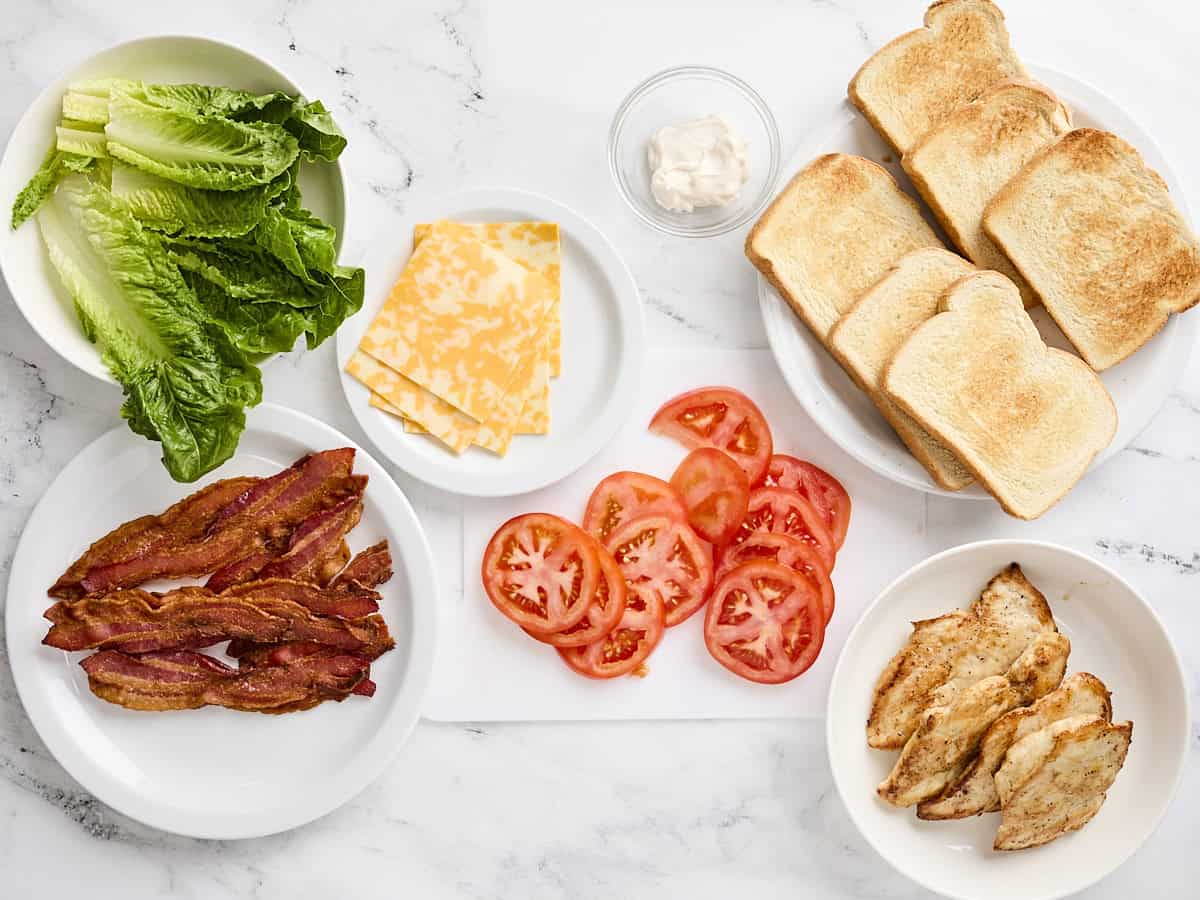 Cooked bacon, romaine lettuce, cheese slices, sliced tomatoes, toasted white bread, and cooked chicken on plates.