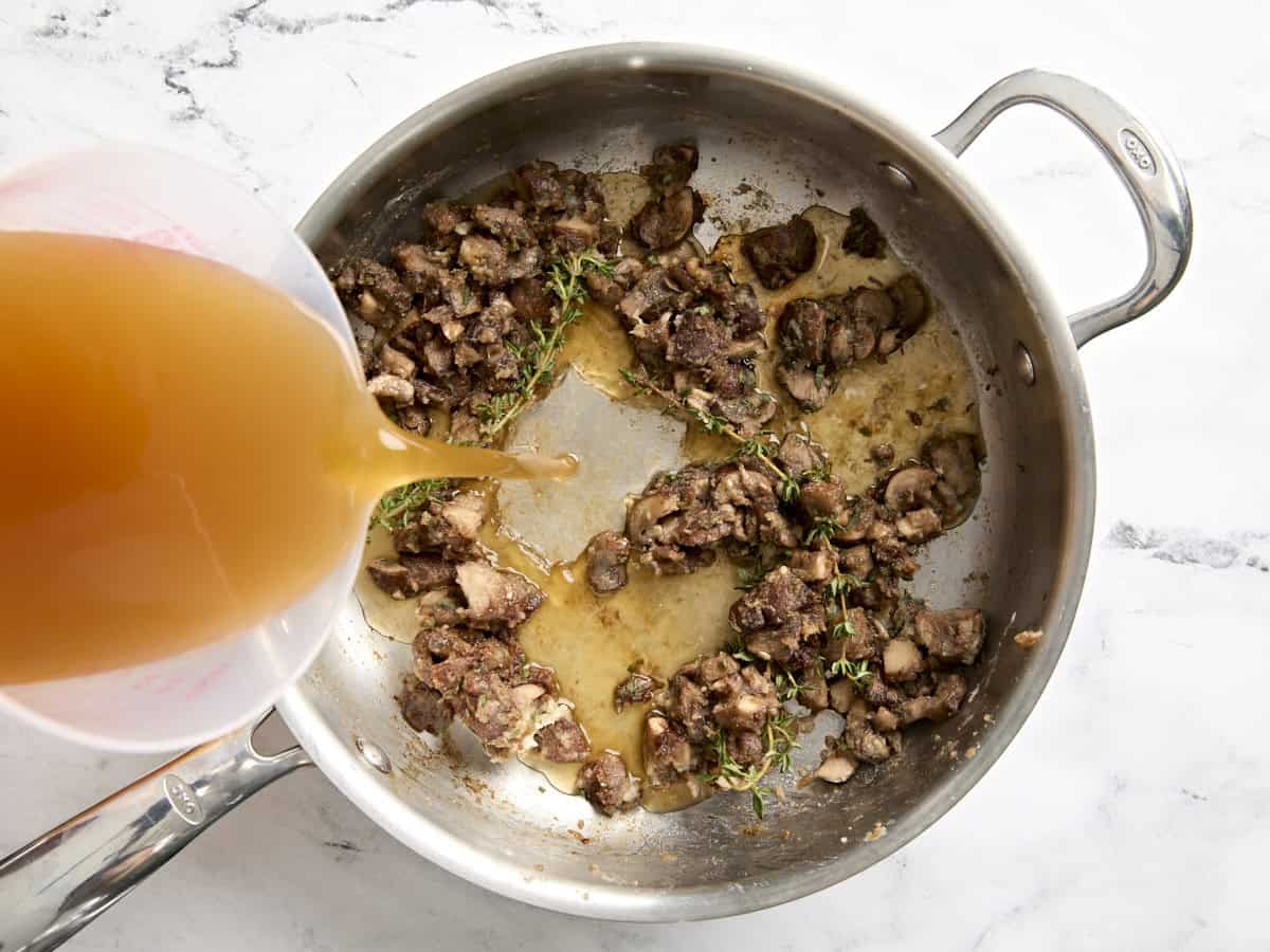 Vegetable broth being poured into a saute pan with mushrooms to make mushroom gravy.