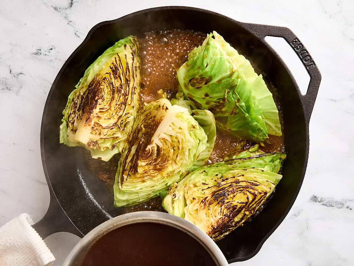 A sauce being poured onto seared cabbage steaks in a cast iron skillet.