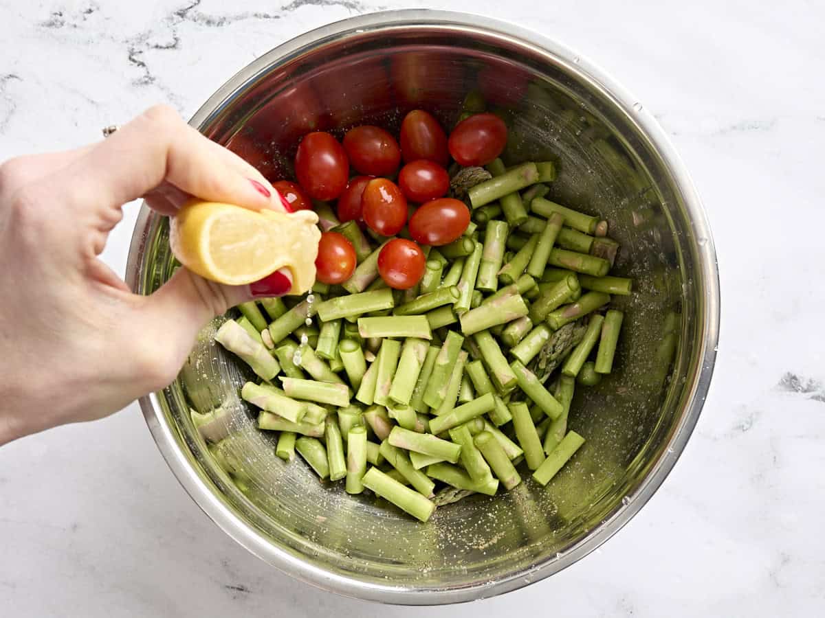 Chopped asparagus and grape tomatoes in a bowl with half a lemon being squeezed over top.