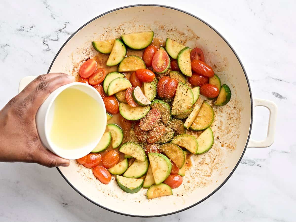 Seasonings and chicken broth added to sauted tomatoes and zucchini in a skillet.