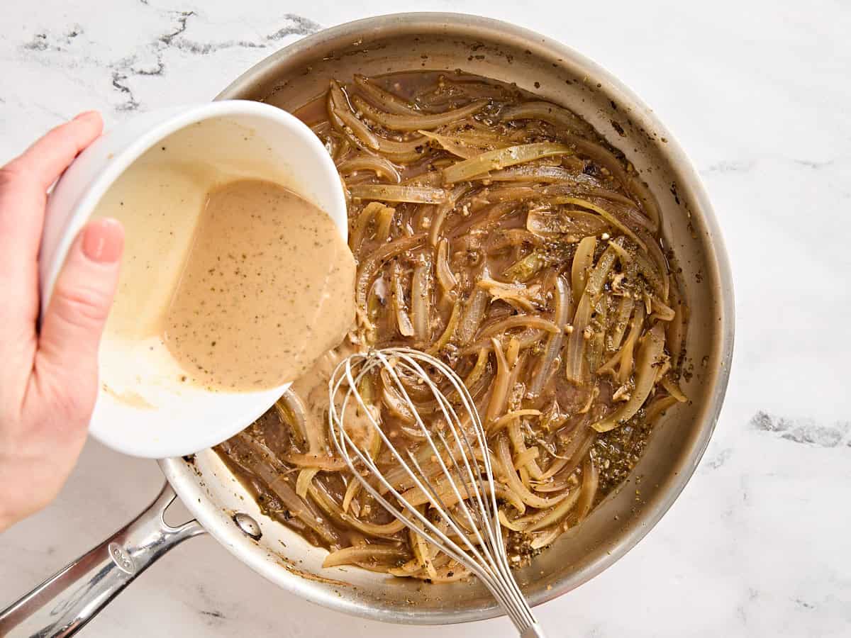 A flour and beef broth slurry being poured into onion gravy in a skillet.