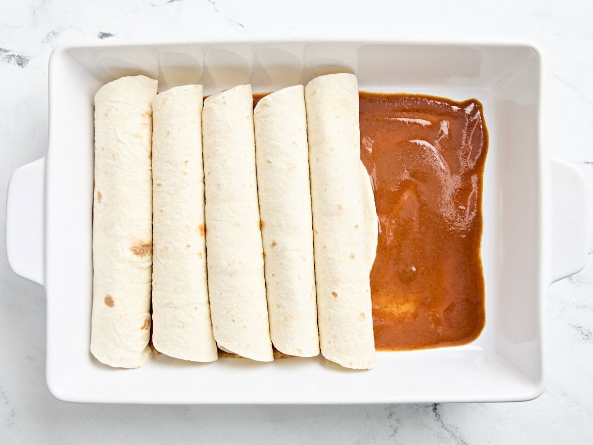 Five chicken enchiladas added to a baking dish with a layer of enchilada sauce underneath.