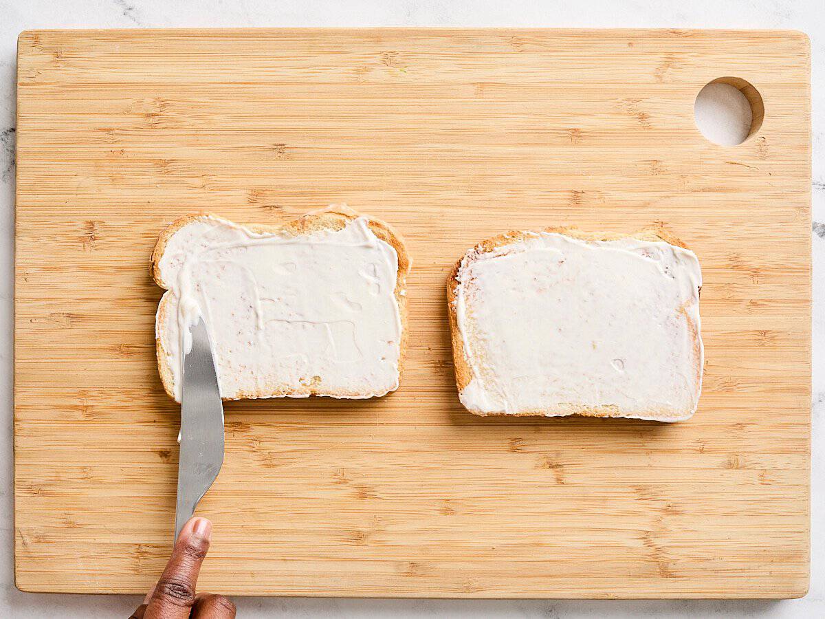 A knife smoothing mayonnaise onto two slices of toast on a wooden cutting board.