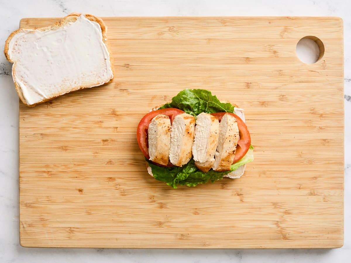 A slice of toast, topped with romaine lettuce, tomato, and chicken on a wooden cutting board.