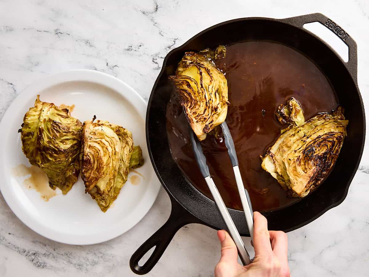 Kitchen tongs removing cabbage steaks from a cast iron skillet to a plate.