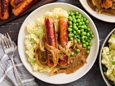 Overhead close up of bangers and mash on a plate with onion gravy and peas.