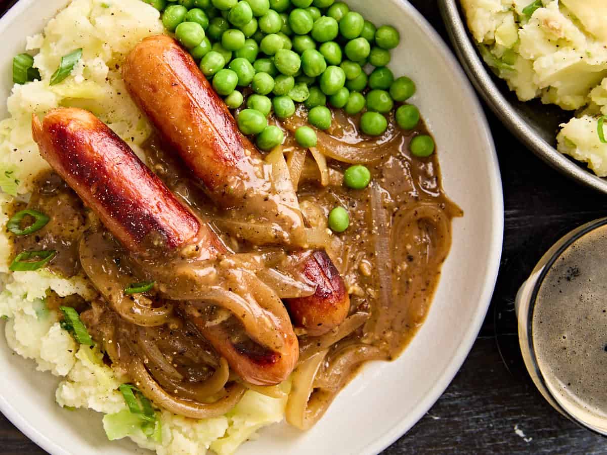 Overhead close up of bangers and mash on a plate with onion gravy and peas.