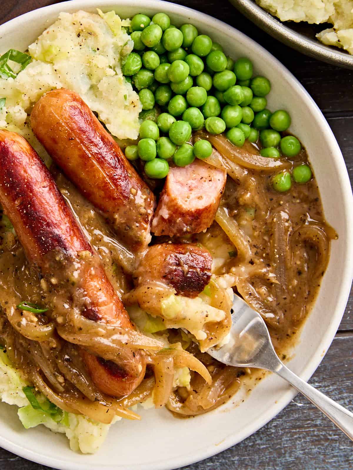 Overhead close up of bangers and mash on a plate with onion gravy, peas, and a fork.