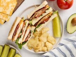 Overhead view of a chicken club sandwich on a plate with chips and a pickle.