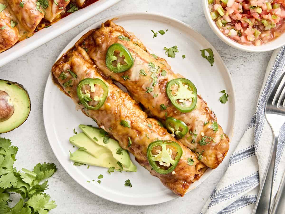 Two chicken enchiladas on a plate with sliced avocado.