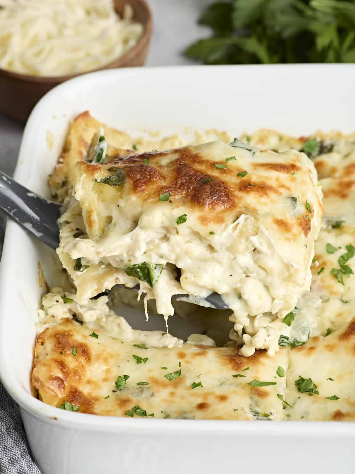 Side view of a slice of white chicken lasagna being lifted from a baking dish. 