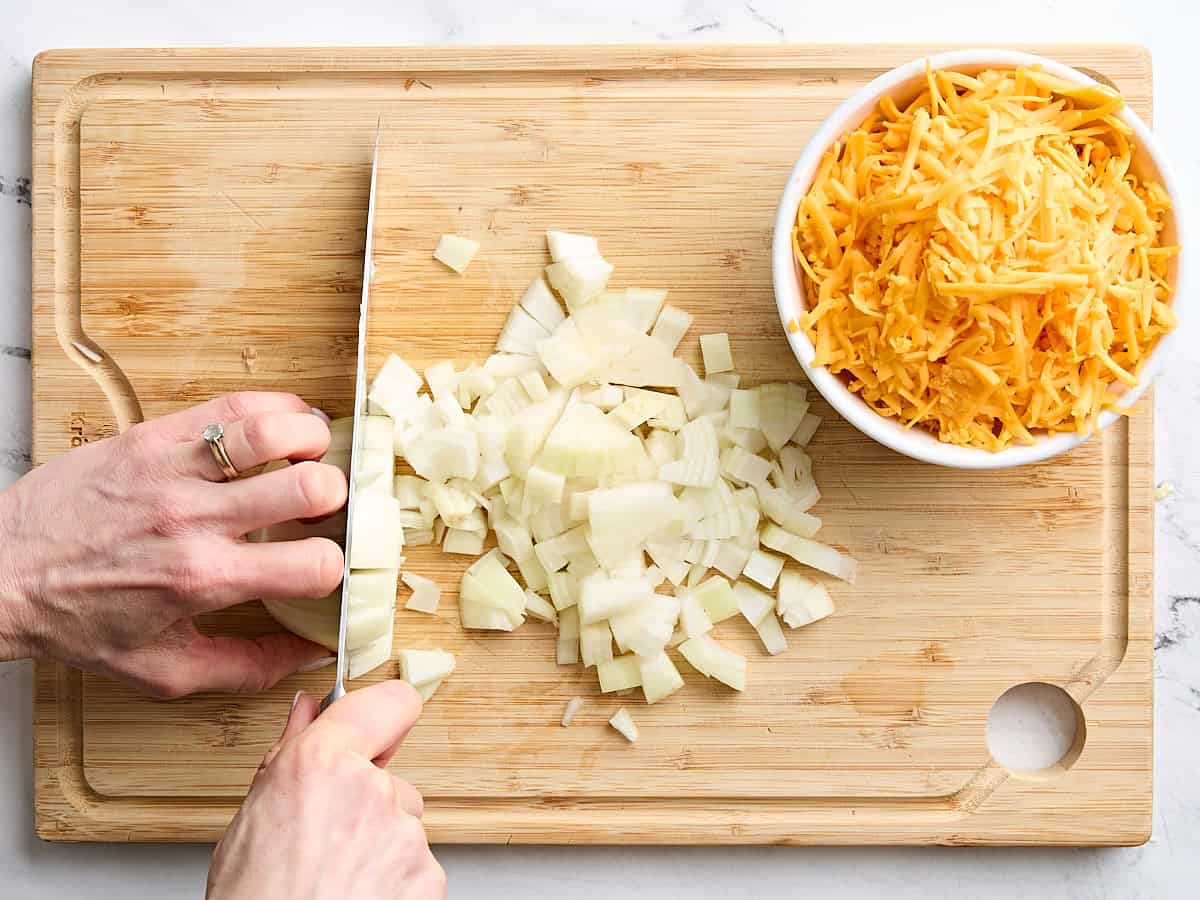 Onion being diced on a wooden cutting board next to a bowl of shredded cheese.