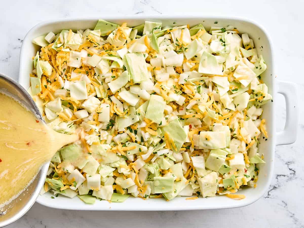 Heavy cream mixture being poured over the assemble cabbage casserole in a casserole dish.