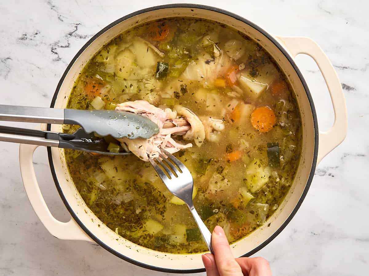 Kitchen tongs holding a chicken breast in stone soup, with a fork shredding the meat from the chicken.